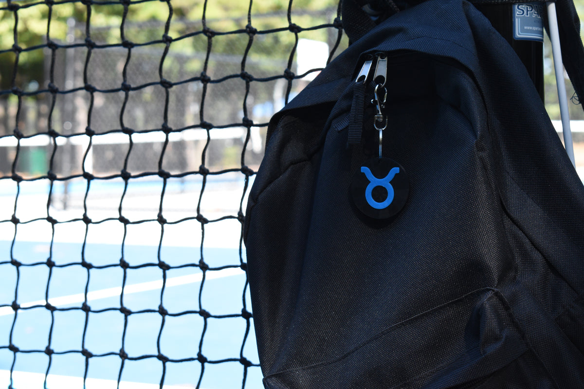 3D printed taurus zodiac keychain clipped to a black backpack zipper outdoors near a tennis court by LayerPlay