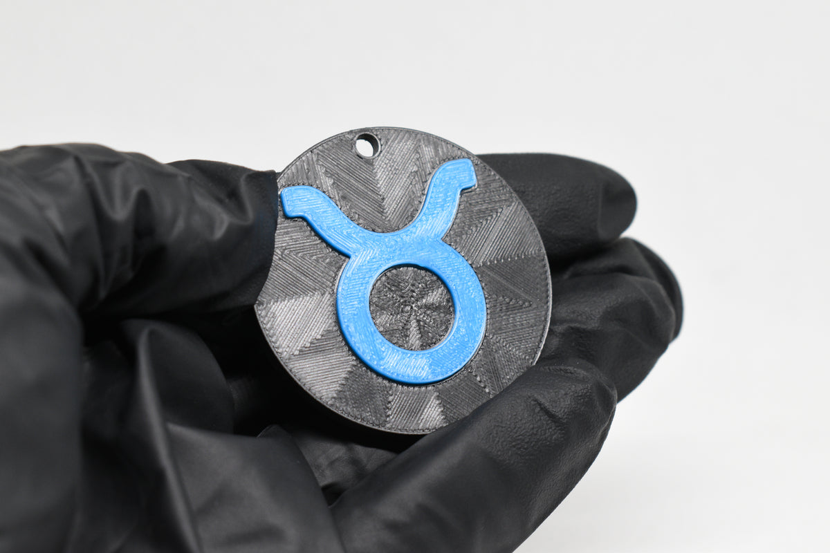 Hand in black glove holding 3D printed blue taurus zodiac symbol keychain front view detail by LayerPlay