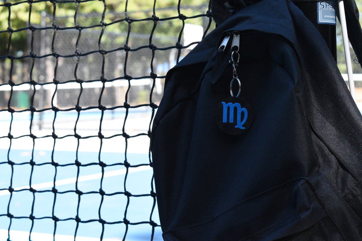 3D printed virgo zodiac keychain clipped to a black backpack zipper outdoors near a tennis court by LayerPlay